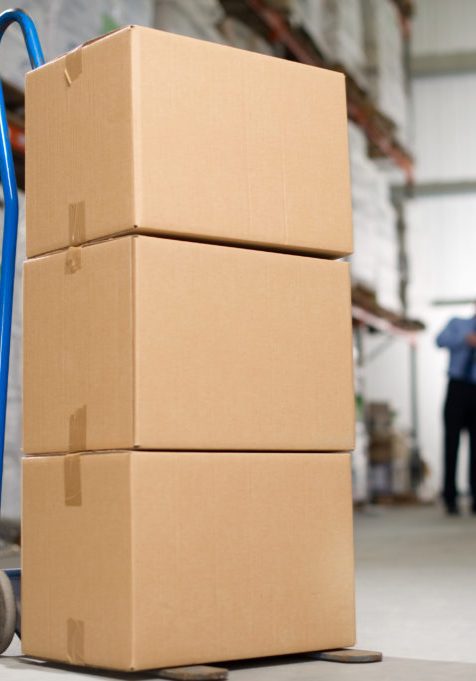 Stack of cardboard boxes sit on a hand truck as two warehouse employees work in the background.